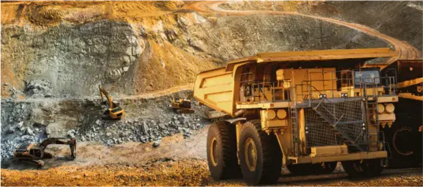 A large yellow mining truck on rocky ground near a pit wall, with smaller vehicles and equipment in the background under warm sunlight.